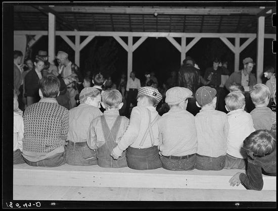 The image depicts a group of young boys sitting on what appears to be a platform or elevated surface. They are dressed in casual, possibly makeshift clothing indicative of the time period and conditions they may have been living under as migrants. The setting seems informal with an outdoor structure overhead that provides partial shelter.

Each boy has their own unique style: some wear suspenders over striped shirts paired with trousers; others sport caps or hats like houndstooth patterns on a couple's headpieces, while another wears what looks to be a cap in the shape of the American flag. One child sits apart from his peers and appears disengaged.

The background is filled with other people who are standing and interacting, suggesting that this might have been during an event or gathering. The atmosphere seems lively yet somber due to the children's expressions which range from neutral to mildly distressed.

There is handwritten text on one side of the photo indicating it as a historical document: "MIGRANTS WAITING FOR SIGNAL TO COME FOR REFRESHMENTS" and additional context given by caption stating, "Halloween party at Shafter camp for migrants, California." The photograph serves not only as an artistic representation but also as a historical snapshot capturing a moment in the lives of these young boys.