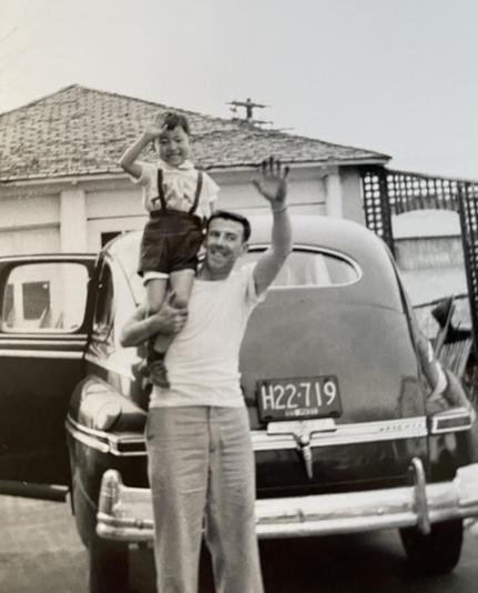 A man is holding a young boy on his shoulders, both smiling and waving. They are standing next to a vintage car in front of a house with a tiled roof. The image is black and white, capturing a joyful moment between the two.