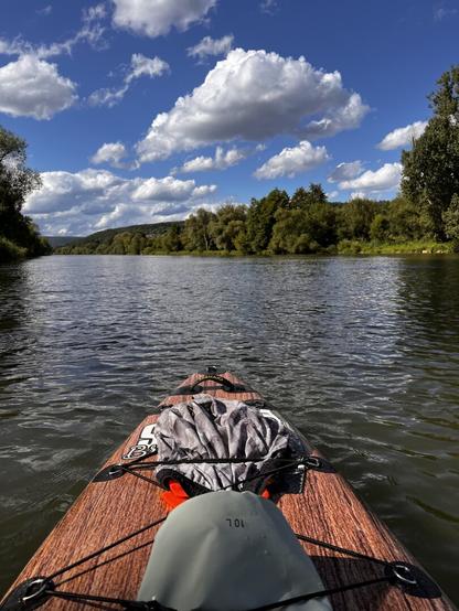 A riverside picture showing the front of a Stand Up Paddleboard. (Very similar to a Surf Board)
There is a dry bag and some clothes straped on to.

The picture was taken in Germany while floating down the river „Main“. It shows parts of the river shore.
No other people are seen. The sunny, warm weather makes it look welcoming and inviting.
A couple „puffy“ clouds can be seen in the otherwise blue, clear sky.