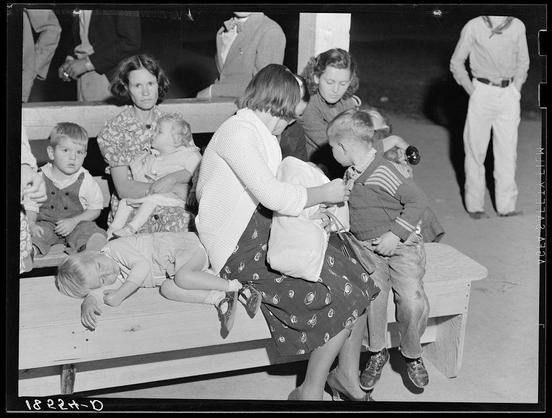 The image is a black and white photograph depicting an outdoor scene at what appears to be a public gathering or event. In the foreground, several women are seated on benches with children in their laps or nearby. One woman is tending to another child who has fallen asleep on one of the seats. The expressions range from concern among some mothers to tranquility and exhaustion amongst others.

The background shows more people sitting around a large gathering area, suggesting that this might be an outdoor fair or community event. Some individuals are engaged in conversation while others appear relaxed but tired.

The attire of the subjects suggests mid-20th-century American fashion with women wearing skirts and cardigans and children dressed in casual clothes like sweaters and jeans. The clothing style indicates a time period consistent with early to mid-1940s America, reflecting modest and practical choices for family outings or community gatherings.

The setting seems informal yet communal, perhaps at an event organized by the local migrant camp where people from different backgrounds come together. The presence of children sleeping on benches hints that this might be a long day's activity leading to fatigue among both adults and young ones alike.

This photograph captures a moment in time reflecting life within communities like those found during the Great Depression or World War II era, particularly with regard to migr [...]