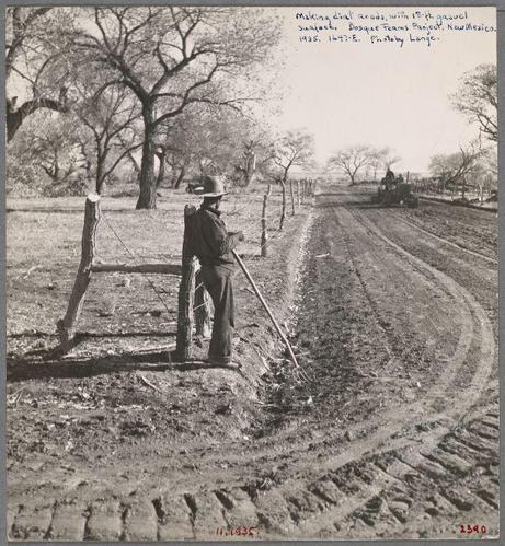 The image is a black and white photograph depicting an outdoor scene from the early 1900s. It shows a person, presumably male due to their attire and build, standing beside a wooden fence on what appears to be a dirt road or path with tire tracks indicating it's used for vehicular travel.

He wears a dark-colored outfit including trousers and jacket, along with boots suitable for outdoor work. On his head is a wide-brimmed hat providing protection from the sun—a common accessory in rural settings during that era.

In front of him lies an old-fashioned rake or hoe which he seems to be either using on or resting against, suggesting agricultural activity possibly related to land preparation or maintenance. His posture indicates contemplation or pause as he gazes into a distance beyond his immediate vicinity.

The surrounding environment features barren trees and bushes indicative of arid conditions common in the American Southwest during early 20th-century rural life. The background shows another vehicle—a tractor with large wheels—indicating ongoing work on improving infrastructure, consistent with historical records that mention projects like "Bosque Farms Project."

There's handwriting at the top left corner which seems to be a description or note about this scene: 'Making diet roads; 18 ft gravel surface,' and references other places such as Bosque Teams Farm, New Mexico. The date on the photo is identified by two red s [...]