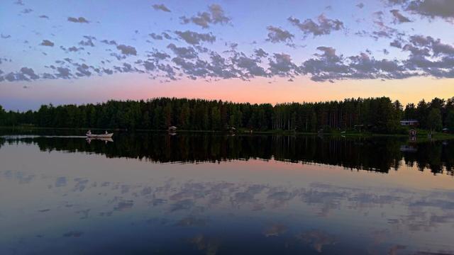 This image captures a serene lake scene at dusk. The water reflects the vibrant colors of the sky, which transitions from soft blues to warm pinks. Fluffy clouds drift across the sky, and the surrounding forest is lush with greenery. A small boat is visible on the lake, adding a sense of tranquility to the landscape. Overall, it conveys a peaceful and picturesque natural setting.