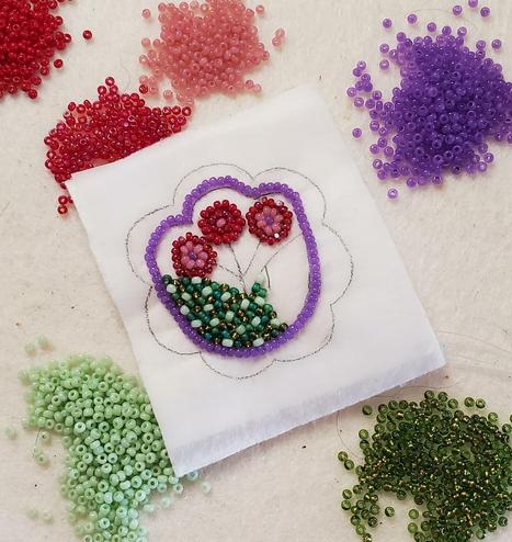 an in-progress beading project made by kiamichi with a floral design surrounded by piles of beads. it has a tulip shaped outline in purple. it's interior has several different shades of green, appearing speckled, with 3 pink and red stamen in the center