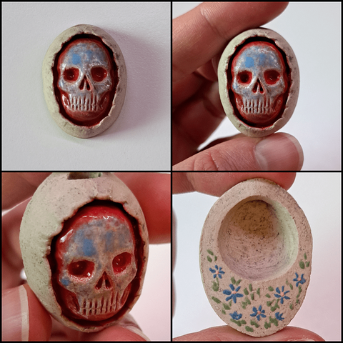 A stoneware clay skull pendant, shown by itself, held, held at an angle and then the back of the pendant. It's coloured by glaze, a blue speckled skull with red around it and in the sockets, surrounded by uncoloured sand-coloured clay. On the back small blue flowers are painted.