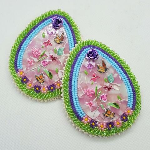 a pair of nature themed, teardrop shaped, beaded earrings made by kiamichi. the centerpiece is a pink enamel with roses, flowers, and monarch butterflies painted on with a metal violet rose at the top. underneath are pink and purple beaded flowers with two rows of light blue and one row or purple beads. all surrounded by translucent vibrant light green beads