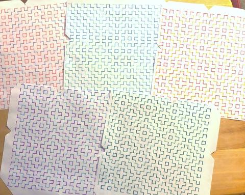 several unfolded handmade envelopes laid out on a table, each is covered in a geometric design made up multi colored horizontal and vertical dashed lines. each envelope's pattern and colors are different.