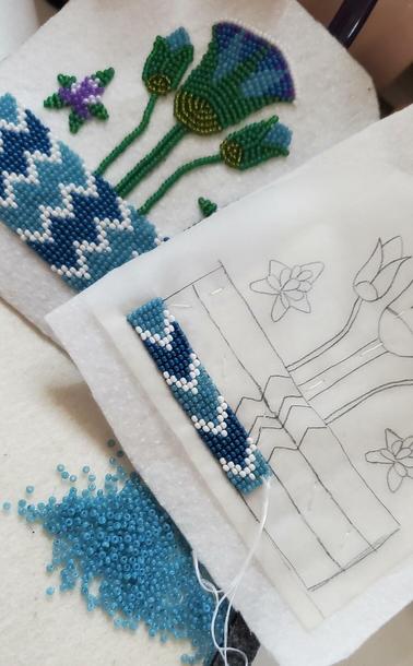 an in-progress beading project by kiamichi of an Egyptian design made with seed beads. the color scheme is teal, blue, white, green, and purple. it depicts an Egyptian lotus coming out of the Nile with some decorative blooming stars next to it