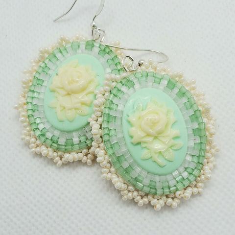 a pair of elegant beaded earrings made by kiamichi. the centerpiece is a cream colored rose on a pastel green background. the surrounding beads are light green and clear and pearlescent to accentuate