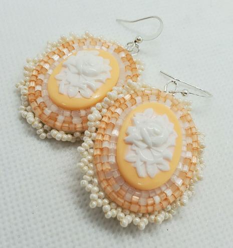 an elegant pair of beaded earrings made by kiamichi. the centerpiece is a cream colored rose on a pastel orange background. its surrounded by clear, shimmery pale orange, and pearlescent beads to accentuate it
