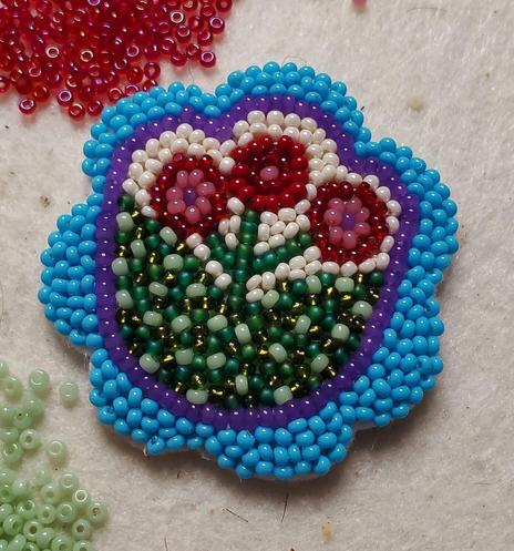 a finished beaded panel intended for earrings by kia of a flower with a light blue border, a purple tulip-shaped outline, and red and pink stamens, filled with many colors of green and cream colored beads. there are two piles of beads nearby in red and green