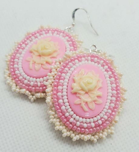 an elegant pair of beaded earrings made by kiamichi. the centerpiece is a cream colored rose on a light pink background surrounded by clear, coral, and pearlescent beads to accentuate.