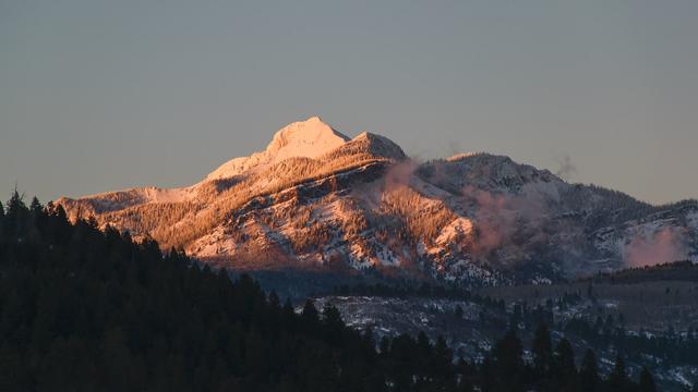 A majestic mountain peak dusted with snow, bathed in the warm glow of sunset, with a forested foreground and a clear sky above.