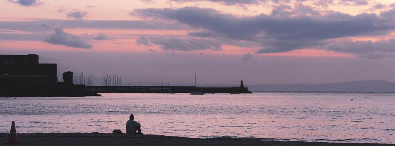 The image depicts a tranquil beach scene at sunset or dusk. There is a person sitting by the shoreline, looking out at the water. The sky is painted in shades of purple and pink with scattered clouds. In the distance, structures like a breakwater or pier are visible, along with some silhouetted buildings. A traffic cone is on the sand near the water.