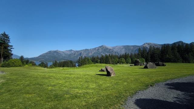 Panoramic image of grassy field in front of a line of tall evergreen trees with mountains across the Columbia River in the distance. Photo taken from the Washington side of the river.