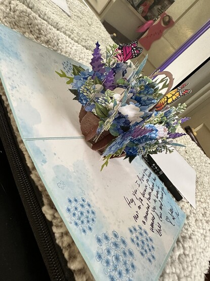 A pop-up paper card with sky, blue and clouds around the edges and a basket of flowers with butterflies in the center.  A short message is written along the side.