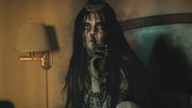 Image of Cara Delevigne as the Enchantress from the Suicide Squad movie holding up a finger to her lips as she crouches on a hotel bed.