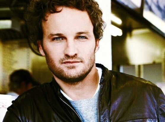 A younger Jason Clarke in maybe a black leather jacket + grey shirt.

His curls are more prominent here, and the slightly stronger stubble or start of a beard is its brown tone here as well. Blue eyes look straight at you.