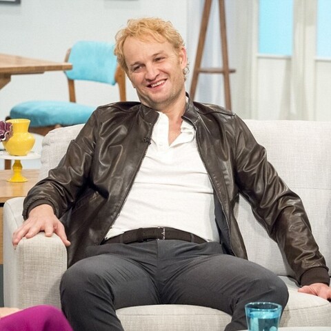 A manspreading dork in badly dyed blonde hair. He played a historical Nazi in a movie some years back and had to go blond for that. It's a deeply unflattering look on him in most pictures.

He's doing a TV appearance I assume, and is leaning back on a pale couch, right hand on the armrest and left palm resting on the sofa cushions. He has a broad, if slightly dorky, smile on his face.

He's dressed in a dark brown leather jacket over a tight white shirt stretched over his upper body. He's wearing tight, darker grey trousers with a dark brown belt. Tight trousers while choosing to manspread and how the picture was taken = I'm not perving on him or that lap of his, but it's presented as if a "See if you can undo that belt - possibly with your teeth" perving challenge as well. The female gaze.