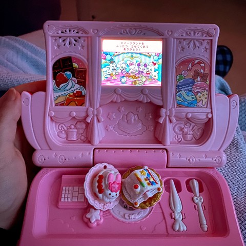 My hand holding a large pink clamshell-folding electronic toy designed to look like a patisserie shop. It has a small backlit touchscreen showing the game, which is basically Cooking Mama for kids, plus some other mini games. Two recipe pieces shaped like different cakes are sitting on top of the clamshell’s bottom half, next to a tiny register, whisk and frosting tool.