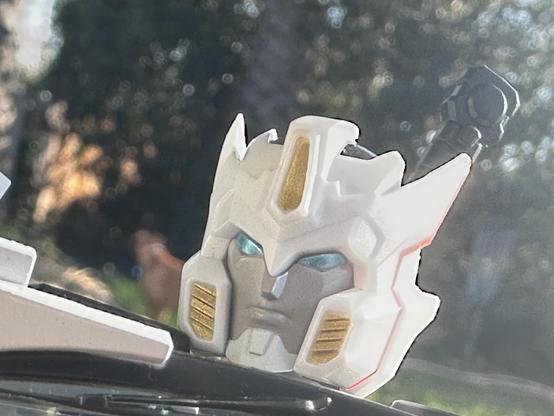 A close-up of a white robot head with blue eyes, featuring gold accents and pointed ears. The background is blurred, suggesting an outdoor setting.