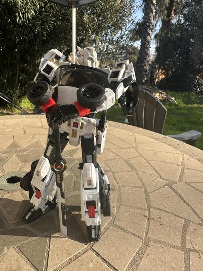 A white and black robot figure stands on a stone table. The robot features a sword and has a detailed design, set against a background of greenery and palm trees.