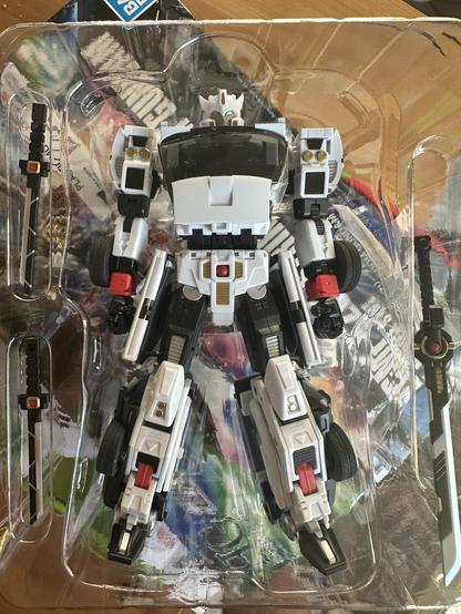 A model figure of a robot is displayed in its packaging. The figure is predominantly white with black accents and features intricate detailing. Two swords are positioned beside the figure in the plastic tray. The background shows a colorful abstract pattern.
