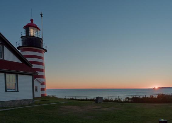 A color landscape photo of a lighthouse, the sea, and the sun rising. The lighthouse and light tower, painted with white and red horizontal stripes, stand on the left side of the frame. A neatly mowed lawn is in the foreground. A flat sea extends out to the sunrise colored horizon. The sun is just beginning to rise from behind a jetty of land on the far right of the frame. The clear is perfectly clear.