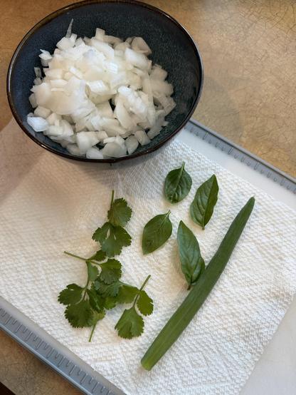 A bowl of diced white onion resting on the edge of a cutting board, on which the following herbs are laid out: basil, cilantro, and green onion.