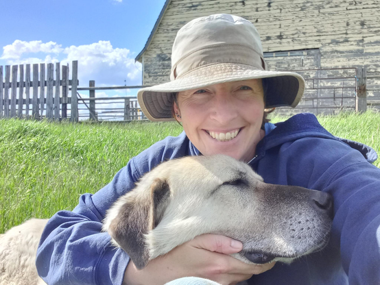 Me, looking right at the camera and smiling, wearing a somewhat grubby farm hoodie and sun hat, with a large yellow dog snuggled up against me, laying across my lap as I sit on the bright green grass.  There is blue sky, puffy clouds, and a large yellow barn in the background.