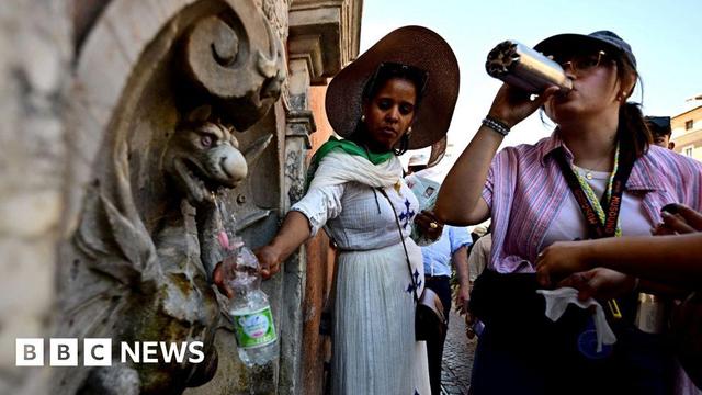 people with wide brimmed hats fill water bottles at a public fountain.