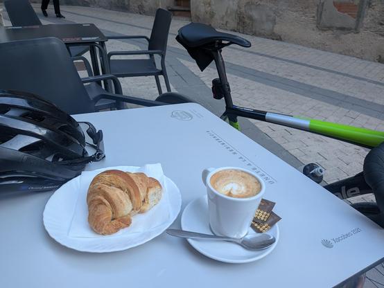 coffee, croissant, and bicycle helmet on a table, with bike leaving against it