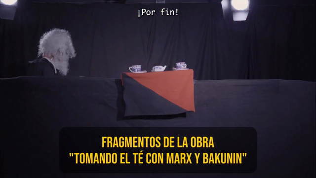 Dos títeres barbones interactúan en un escenario negro con bandera rojinegra.