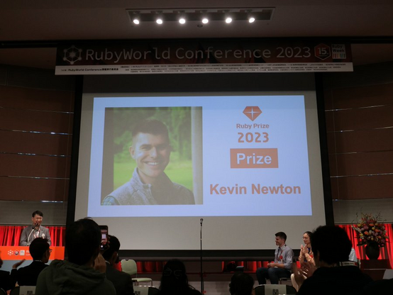 Photo of a conference stage with a large projection screen showing the announcement of the winner of the Ruby Prize 2023