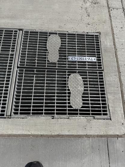 Picture of a grate in a sidewalk in downtown Calgary. The grate has 2 flat plates shaped like footprints welded on top.