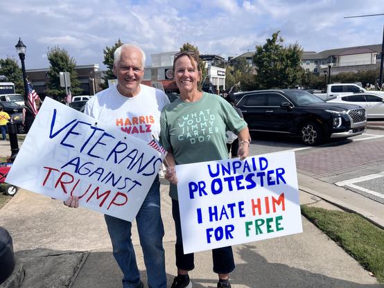 Two people holding signs at No Kings Rally in Smyrna, GA. Woman in Green shirt that says “What Would Jimmy Carter Do”  Holding a sign that says “UNPAID PROTESTER I HATE HIM FOR FREE” Man in white shirt With Dunkin Donuts stylized “HARRIS WALZ” holding sign that says “VETERANS AGAINST TRUMP”