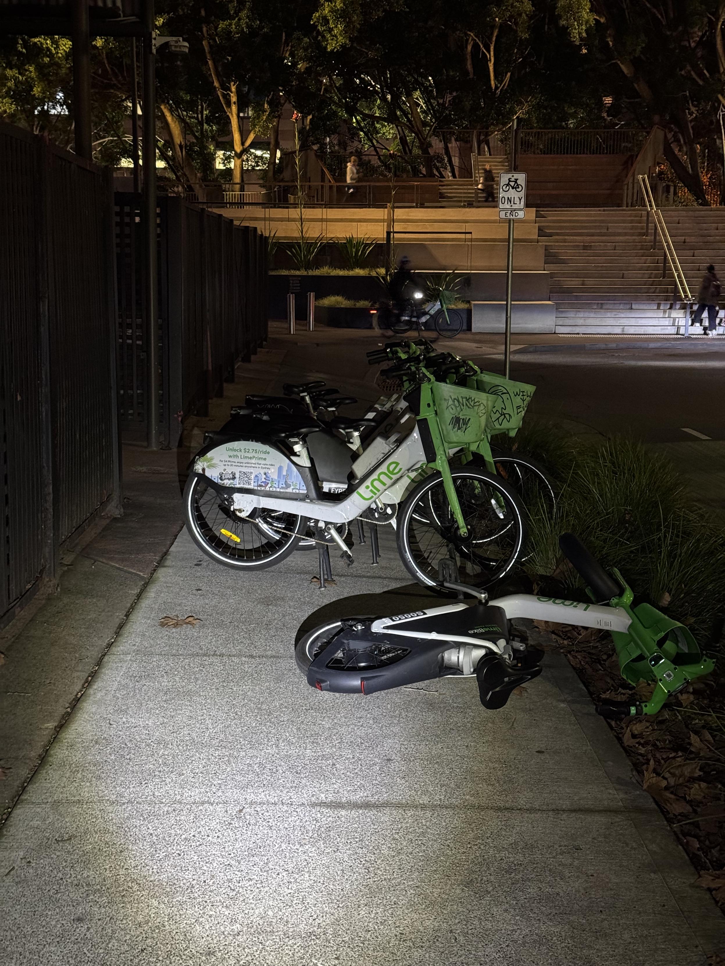 Pavement in Sydney, Australia with six Lime bikes parked sideways across it, leaving only a small gap for people to walk around them. One bike is knocked over on its side.