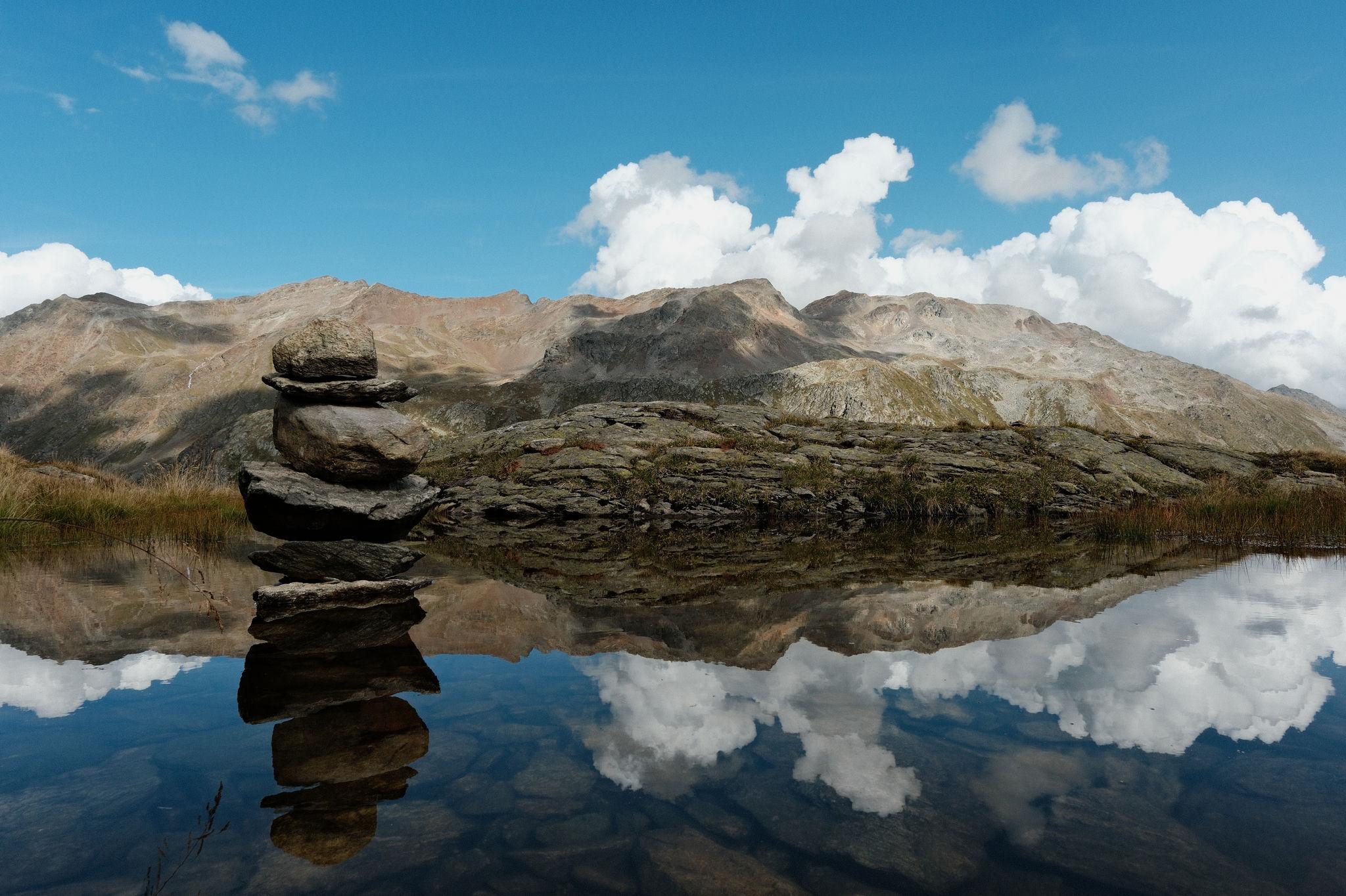 A few small rocks piled up to a tower in a small pond. The tower, sky and the mountains in the back reflect in the pond giving a perfect mirror image.
