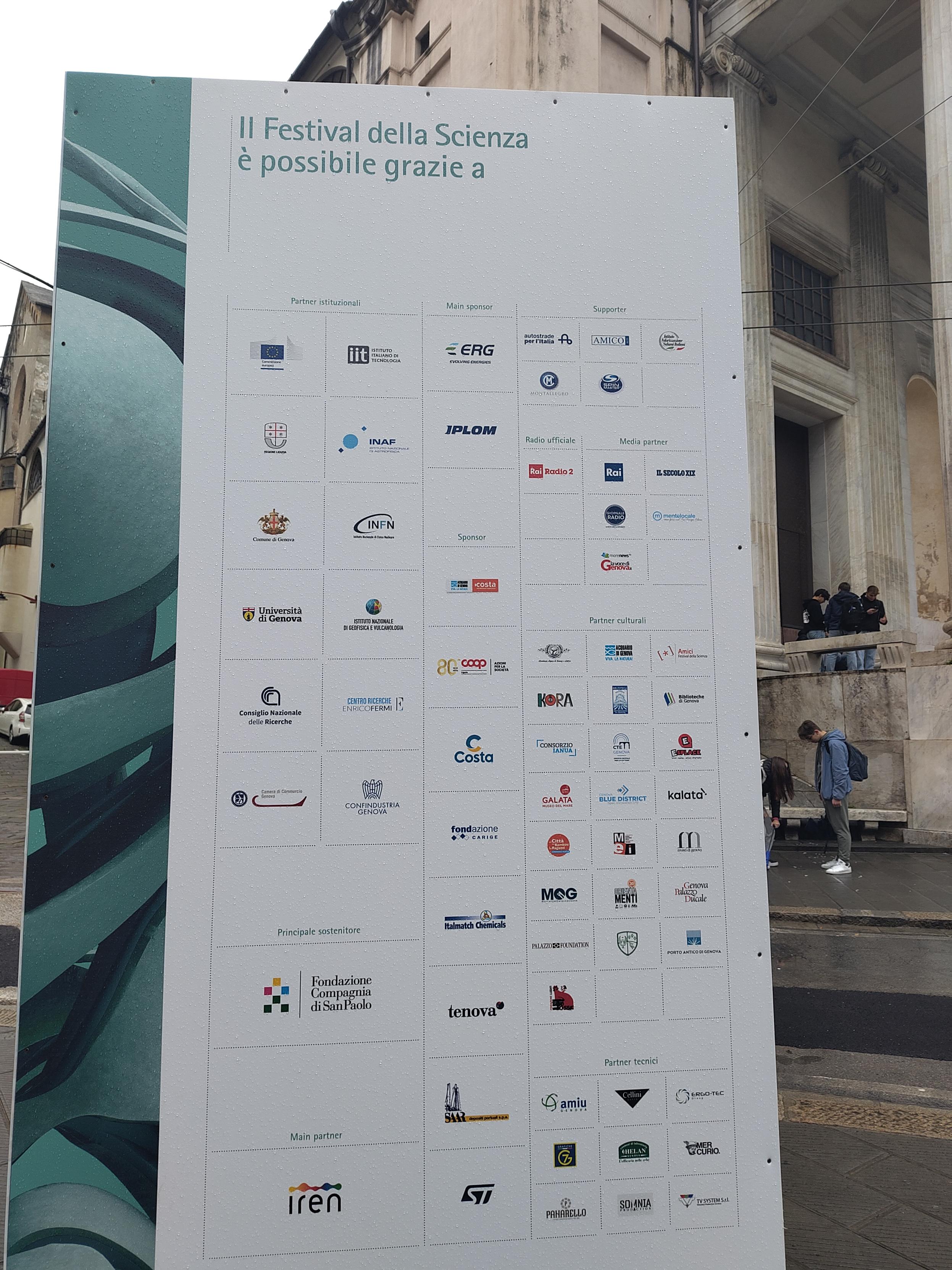 Un grande pannello informativo in piazza della Nunziata a Genova, fotografato in una giornata piovosa.
C'è una lunga lista di logo di enti, aziende e organizzazioni che sostengono il Festival della Scienza. 
I main sponsor sono ERG, azienda petrolifera, e Iplom, raffineria.