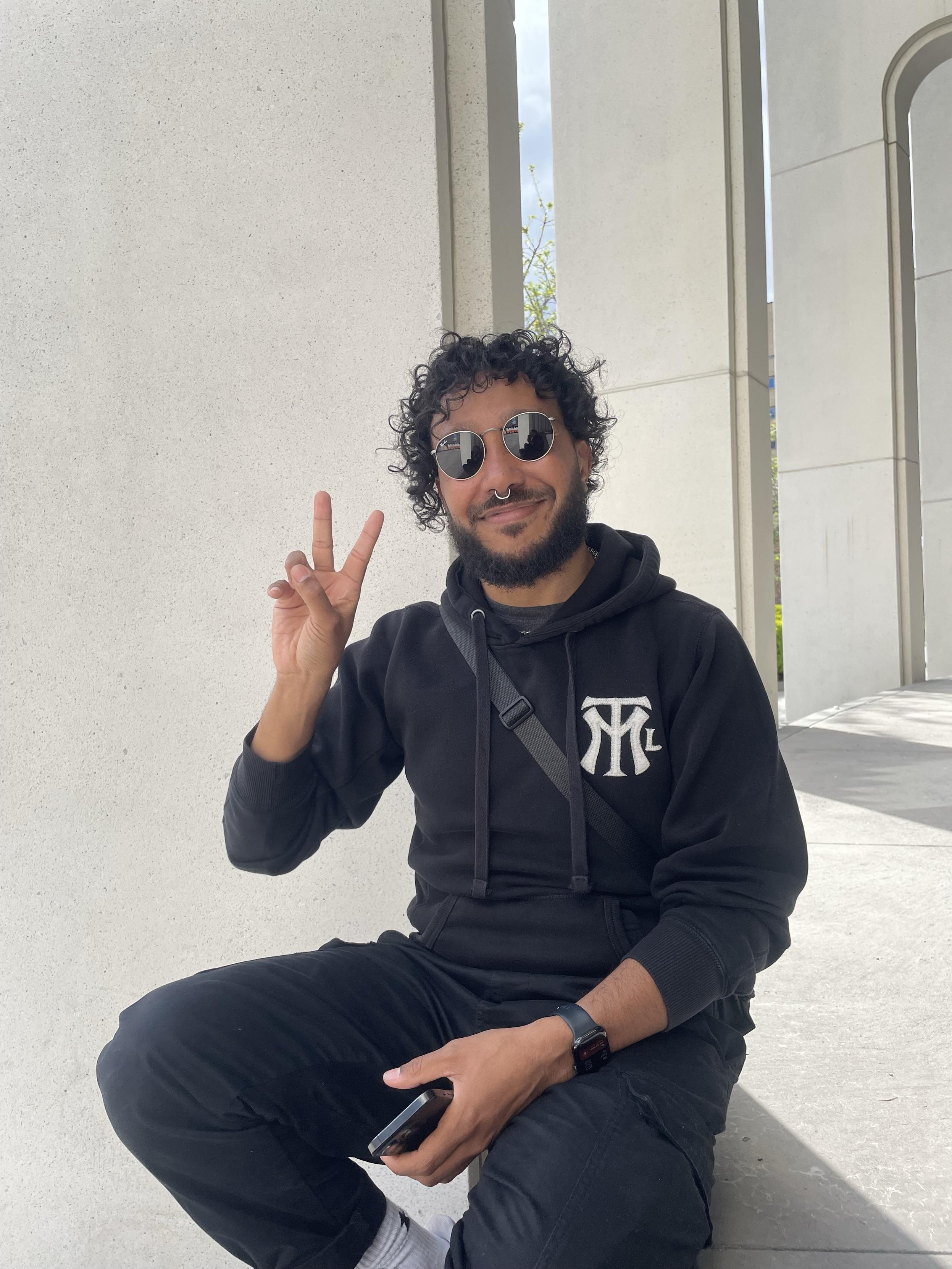 Vincent (that’s me!) sits on concrete steps against a white stone building, wearing an all-black hoodie with a monogram logo from Artgang Montréal that looks like the NY Yankees logo but with MTL instead, black cargo pants, and round sunglasses. They have curly dark hair, a beard, and a septum ring, and are holding up a peace sign with one hand and a phone in the other. A crossbody bag strap crosses their chest. They are smiling softly at the camera.​​​​​​​​​​​​​​​​