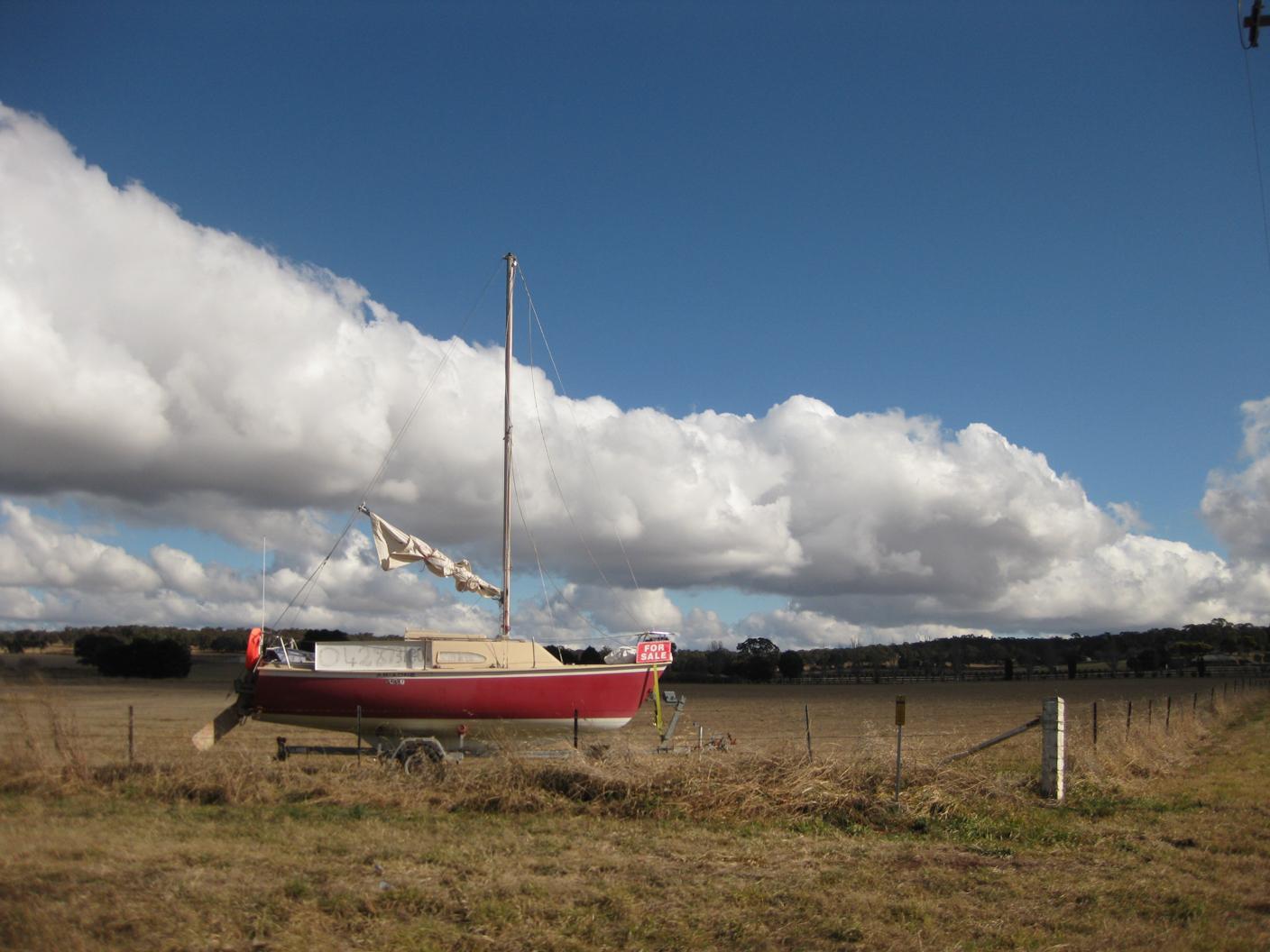 Photograph of a small sailboat on a trailer in a field behind a barbwire fence. There is a FOR SALE sign on the front of the boat. A dramatic arc of low white clouds echo the furled up roll of sail. Rural New South Wales, Australia, 4 July 2011. Photo Cindy Kohtala