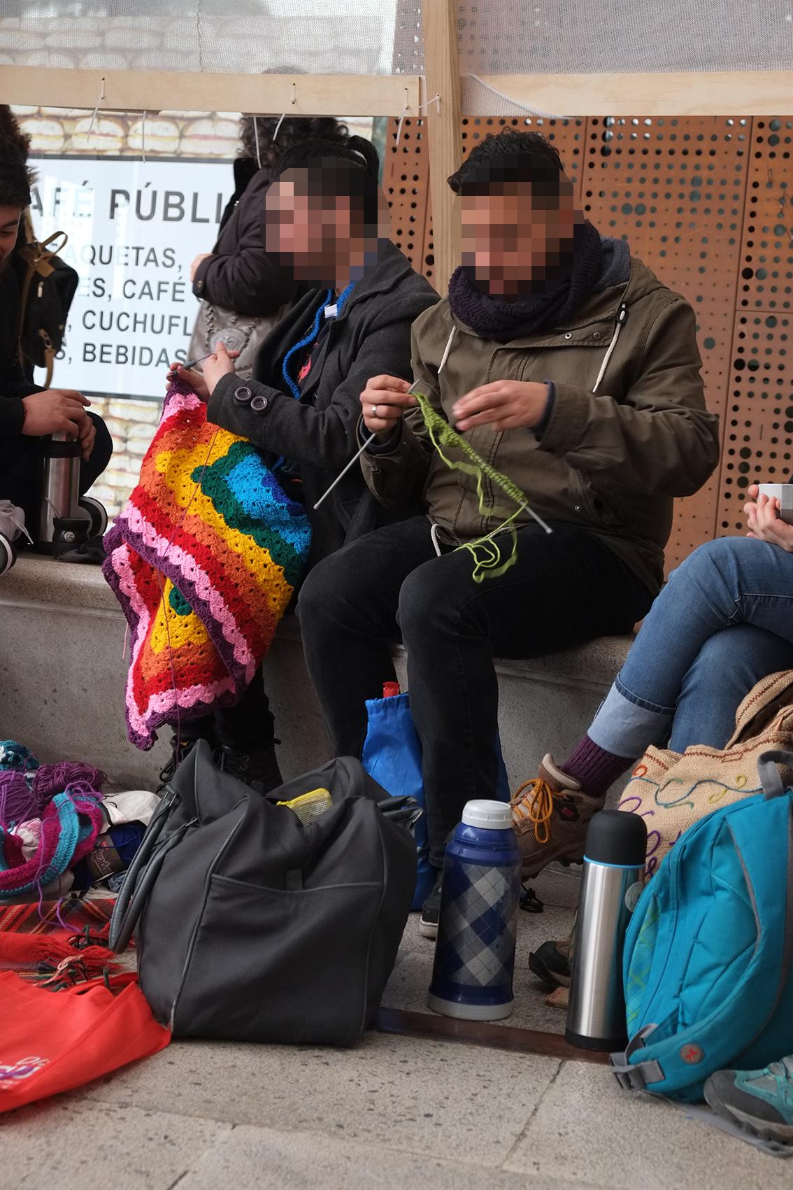 Photo of some people sitting together knitting outdoors in a public space. Their faces are pixelated/anonymized. In the centre two men are knitting, one of them is knitting a large and colourful afghan blanket. There are others beside them out of frame. Coffee thermoses and bags of materials are on the ground. Santiago, Chile, 5 August 2017. Photo Cindy Kohtala