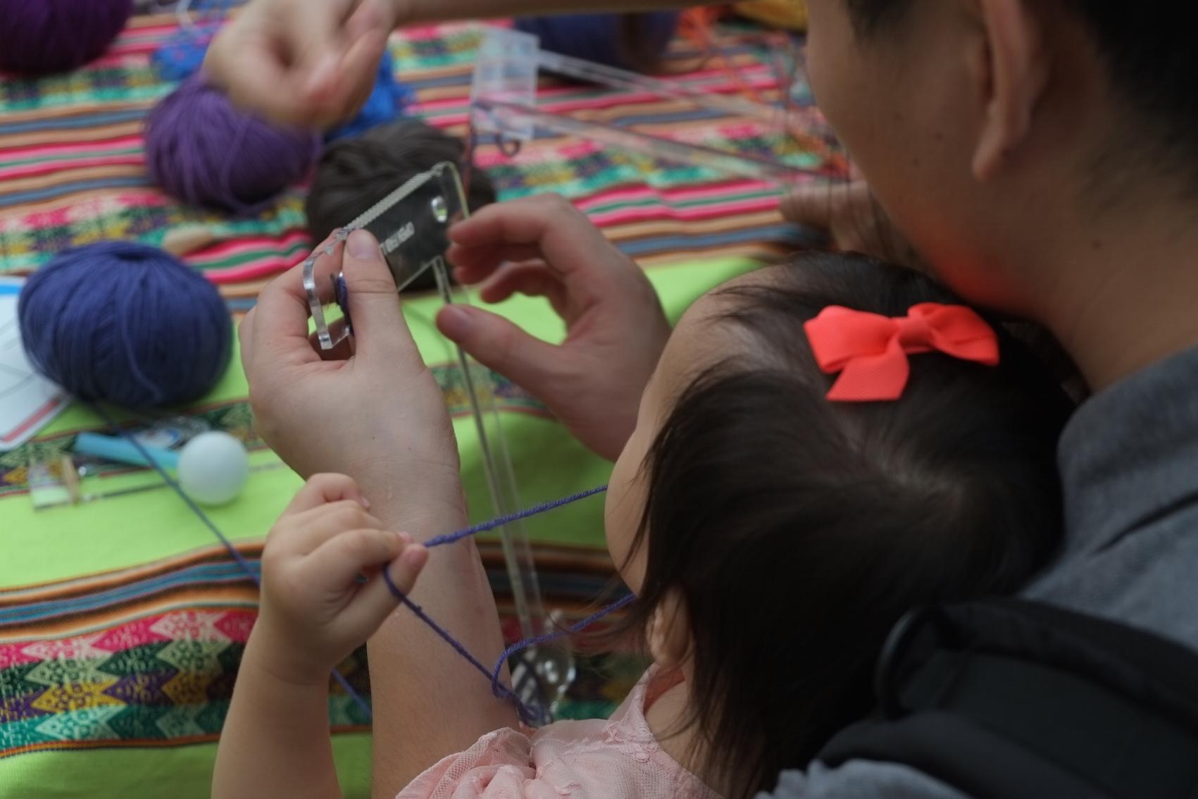Photo of a father and daughter, a young child with a bow in her hair and sitting on his lap, from the back, focusing on their hands. They are working with a small lasercut acrylic loom. The girl is pulling on a thread of blue yarn and the father is holding the loom about to thread it. Balls of different coloured yarn are on a table in front of them. The table is covered with a brightly coloured Peruvian woven textile. FAB12: Fabricating the Future, Shenzhen, China, 13 August 2016. Photo Cindy Kohtala