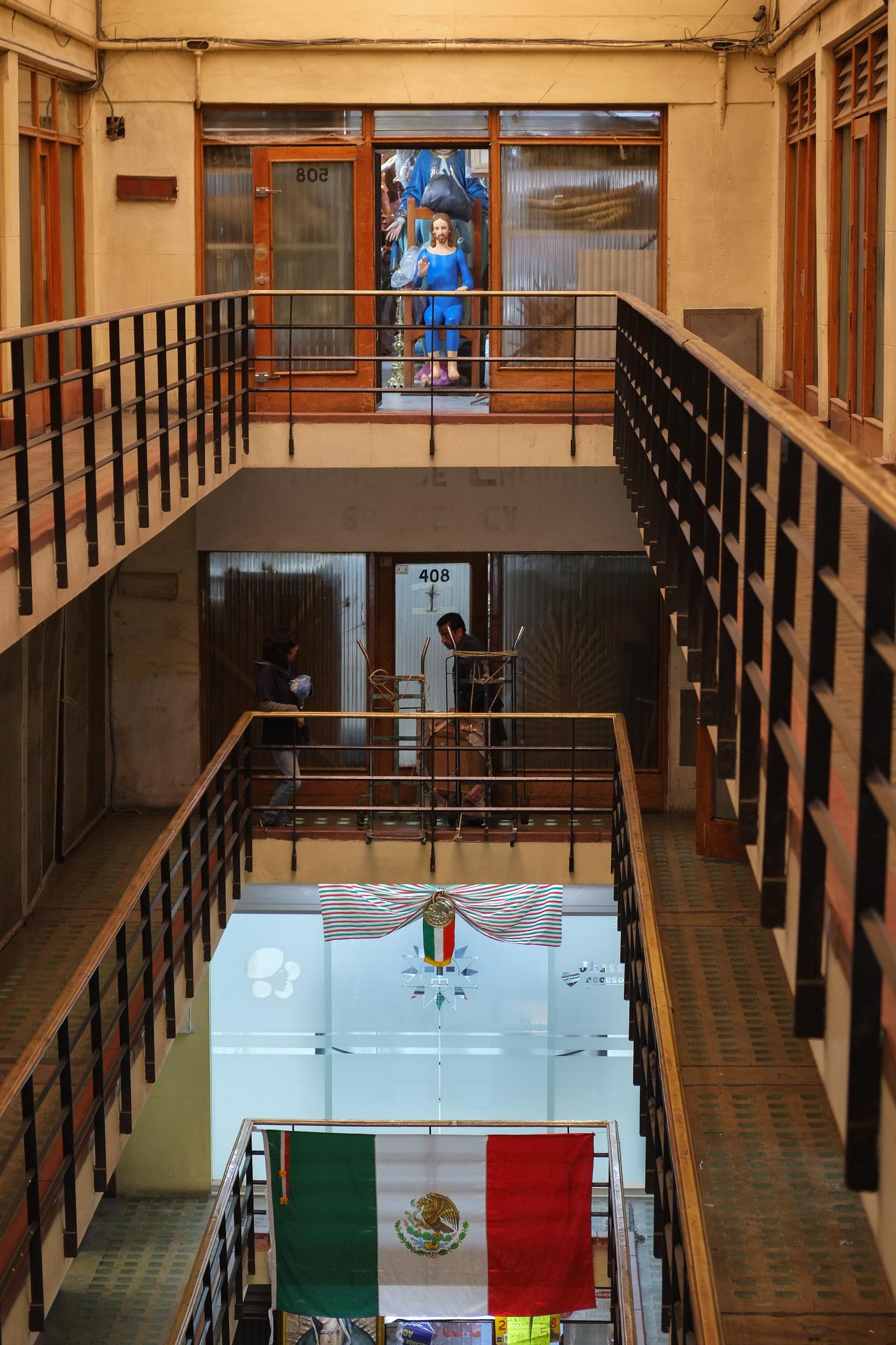 Photo inside a specialized retail building for Catholic church items. The building has a central courtyard inside that extends for the height of the building and railed mezzanine levels around the centre - from which doorways lead to businesses. Two levels down, a large Mexican flag is hanging from the railings. One level down, a delivery person is walking with goods on moving trolleys. At the same level on the other side of the building, there is an open door where a full size statue of Jesus is sitting on a large wooden chair raising one of its hands up, with some plastic wrap behind it. Other religious items are visible behind it. Right behind the main Cathedral at the Zócalo, Mexico City, 12 September 2016. Photo Cindy Kohtala