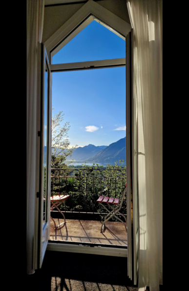View from my hotel room in Ascona of Swiss mountains and a small sliver of the blue Lago Maggiore, framed by a variety of different trees on a bright and sunny day with blue skies and two tiny clouds dotted above the mountains. Two tall glass doors open out onto a small balcony with two red chairs in the foreground. Above the doors is a triangular window.
