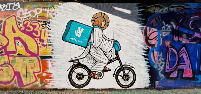 A wall of graffiti - yellow and red letters on the left and purple/ blue ones on the right with the middle being painted over  in white brush brush strokes. A white clothed jesus figure with halo on a bicycle is carrying a deliveroo box as a backpack. World peace is printed underneath the deliveroo logo. Next to the back wheel, some small print reads "Happy Easter G'gow!"