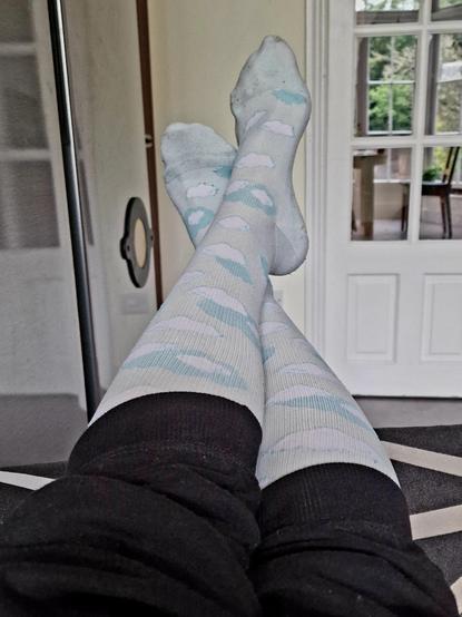 Stretched out feet on a black patterned sofa, left foot crossed over the right, adorned with light blue / turquoise compression socks with cloud pattern