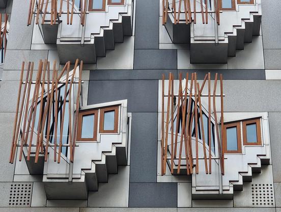 Four individual complex shapes protrude from the facade of the Scottish parliament, two seen in full and two cut off.
Each is comprised of two small rectangular windows with brown wooden frames and a larger bow-shaped one to the left, half hidden behind wooden sticks acting as shading. There are small steps at the bottom right and one large one at the bottom left, behind which a seat is hidden.