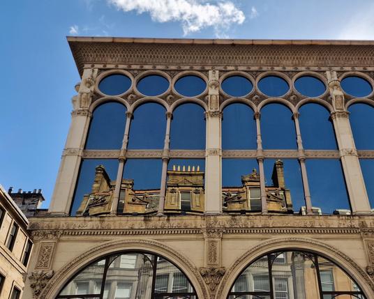 Top floor of the Ca d'Oro building with its elegant façade, thin columns and decorative elements in bright sunshine against blue skies and a tiny cloud above it. A traditional brick building opposite is mirrored towards the bottom of the top floor. It looks as if the brick structure opposite sits within the Ca d'Oro building.
