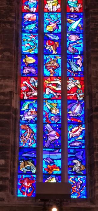Bottom three quarters of a tall stained glass window in the cathedral of Palma de Mallorca, photographed from the inside. It's a modern design with three panels next to each other, from bottom to top four rows of sea animals against blue background, one row with soldiers against red background, then three more rows of sea creatures and fish