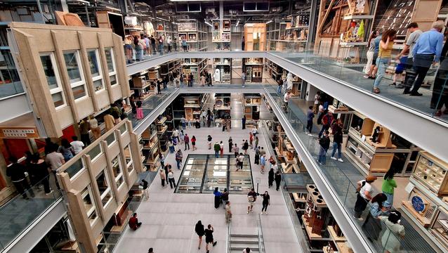 View from the top floor into the main hall of the V&A East Storehouse - a rectangular foyer framed by walkways with all sorts of exhibits on display. Lots of people are dotted around the place